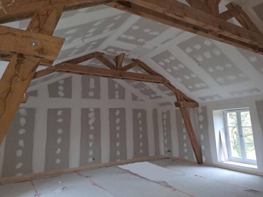 Rénovation de combles avec poutres apparentes et placo Rénovation de combles avec plaques de plâtre et charpente bois apparente dans une pièce lumineuse en travaux.